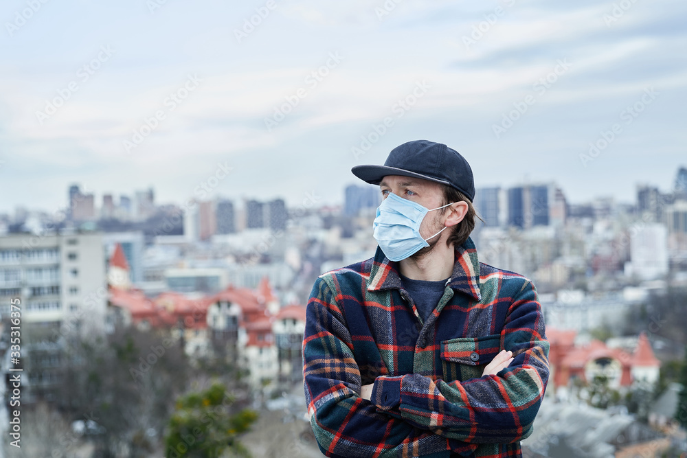 Young European man with a medical face mask on the roof with big city view. An attractive male in medical mask to protect against corona virus infection. Covid-19 or pollution concept