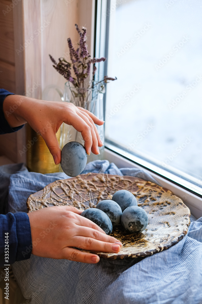 Blue Easter eggs in the hands of a girl near the window. Rustic style, natural materials and textiles, Crystal decanter, ceramic plate in the background. Wooden wall.