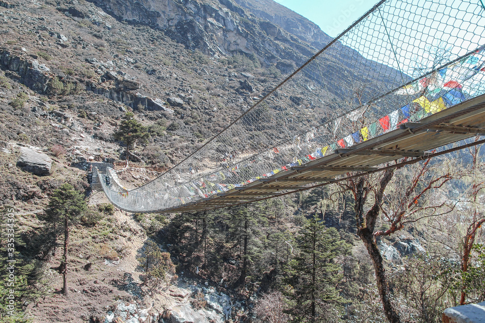 One of many pedestrian suspension bridges with waving prayer flags in Himalayas on the way to Everest Base Camp in Nepal. Clear blue sky. Side view. Theme of trekking in Himalayan mountains. - obrazy, fototapety, plakaty 