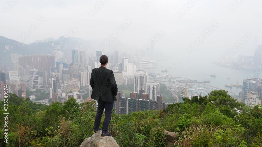 Lonely figure at top of hill, man look to Hong Kong in morning mist from height. Guy wear casual suit, stand at stone surround with green bushes. Urban areal crowded with tall buildings in fog Lonely figure at top of hill, man look to Hong Kong in morning mist from height. Guy wear casual suit, stand at stone surround with green bushes. Urban areal crowded with tall buildings in fog