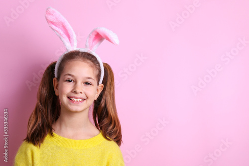 Studio portrait of smiling young girl wearing traditional bunny ears headband for easter. Portrait of brunette female with pigtails in blank yellow sweatshirt. Pink background, close up, copy space.