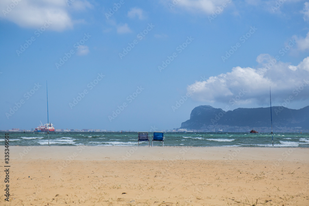 landscape of a golden sand beach, blue sky with bkanca clouds, on the shore there are two empty hammocks and fishing rods, boats in the sea and in the background we see the rock of Gibraltar