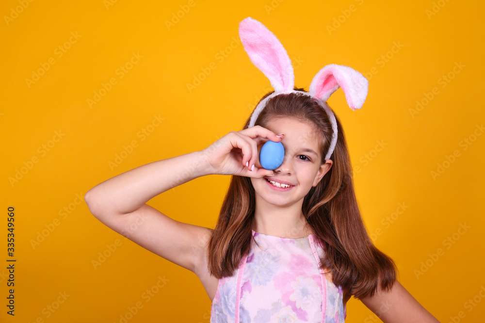 Studio portrait of young girl wearing traditional bunny ears headband for easter, covering her eyes with an egg and smiling. Brunette female with pigtails over yellow background. Close up, copy space.