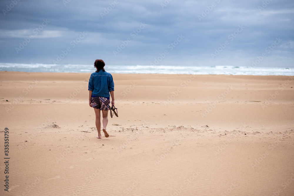 Woman walking alone on empty beach with sandals in her hand