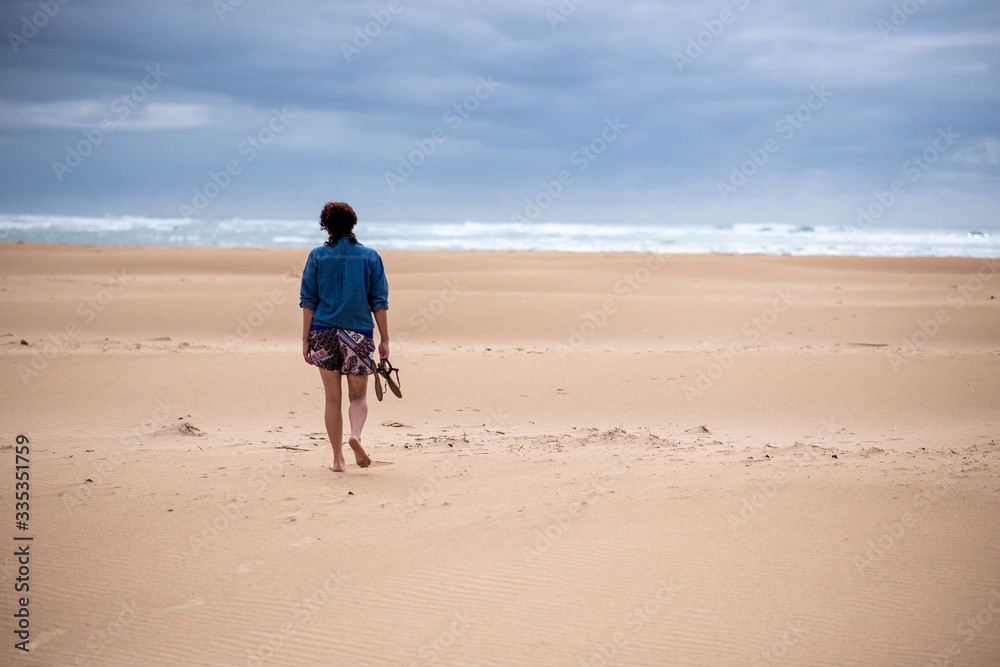 Woman walking alone on empty beach with sandals in her hand Woman walking alone on empty beach with sandals in her hand