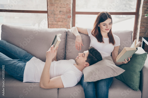 Portrait of his he her she nice attractive lovely focused cheerful cheery couple sitting lying on divan reading interesting book spending time pastime at modern industrial loft style brick interior