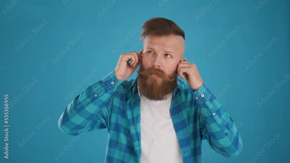 portrait of Caucasian man with beard and mustache in blue shirt, inserts headphones in his ears, listens to music, enjoys, dances and smiles, on blue background close-up 4K frame portrait of Caucasian man with beard and mustache in blue shirt, inserts headphones in his ears, listens to music, enjoys, dances and smiles, on blue background close-up 4K frame