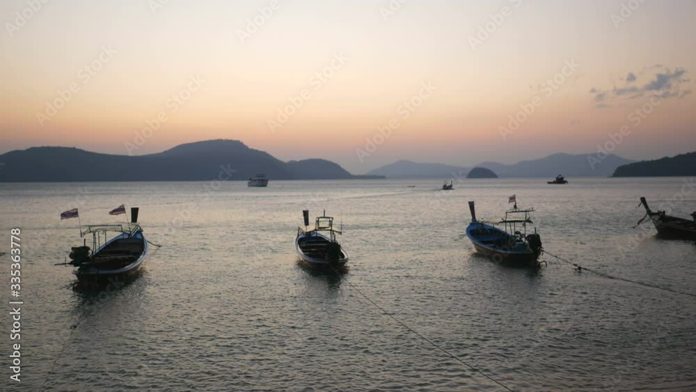 soft water wave breaking to shore, moving thai flag on parking long tail boat tied with long rope, sailing boat background, silhouette mountain and island, in twilight orange color sky evening time soft water wave breaking to shore, moving thai flag on parking long tail boat tied with long rope, sailing boat background, silhouette mountain and island, in twilight orange color sky evening time
