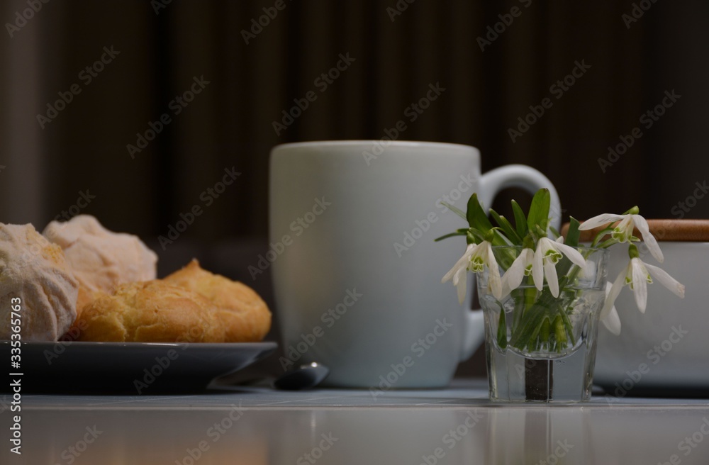 Tea and plate with eclairs and marshmallows on a transparent table. Vase with snowdrops. Tea and plate with eclairs and marshmallows on a transparent table. Vase with snowdrops.