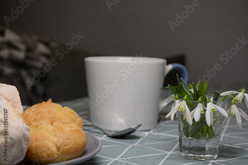 Morning breakfast in the home kitchen. Tea and plate with eclairs and marshmallows on a tablecloth with a triangular pattern. Vase with snowdrops. Blue-green-gray tone