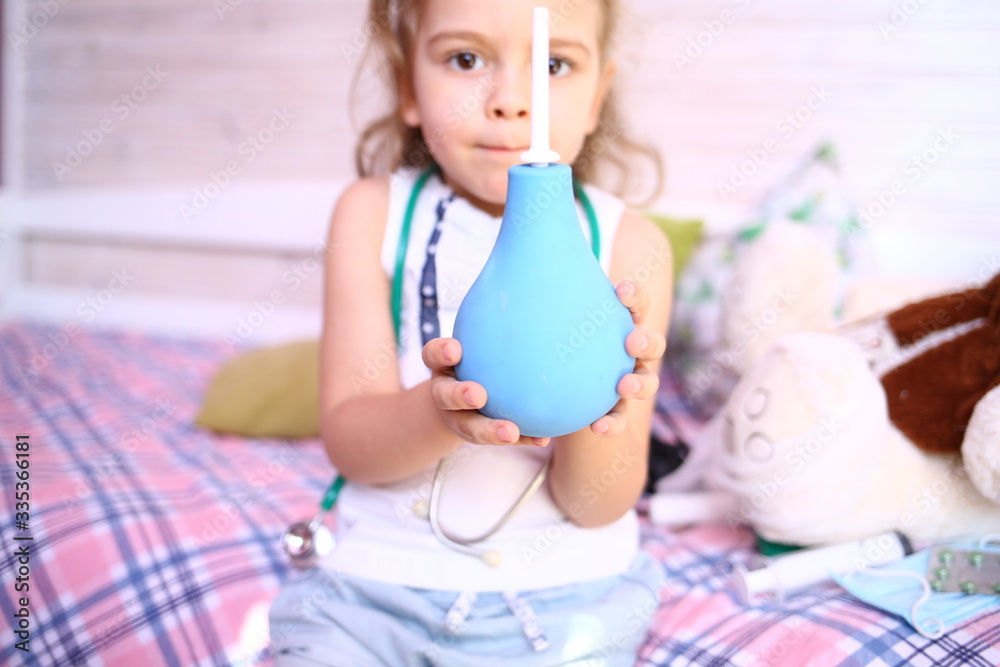 Caucasian little girl with stethoscope at home playing doctor with big blue enema to treat a bear. Little therapist. Coronavirus 2019-ncov. Home insulat Caucasian little girl with stethoscope at home playing doctor with big blue enema to treat a bear. Little therapist. Coronavirus 2019-ncov. Home insulat