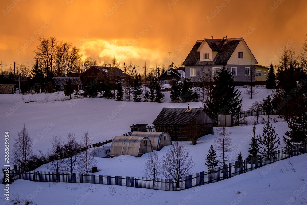 Beautiful winter landscape with a small village on a hill against the backdrop of a sunny sunset in the cloudy sky. Russia, Arkhangelsk region