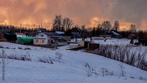 Beautiful winter landscape with a small village on a hill against the backdrop of a sunny sunset in the cloudy sky. Russia, Arkhangelsk region