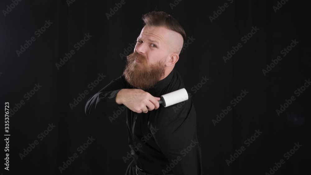 portrait of a Caucasian man with beard and mustache in black shirt and jeans cleaning clothes with roller from dust and particles black background close up 4K frame portrait of a Caucasian man with beard and mustache in black shirt and jeans cleaning clothes with roller from dust and particles black background close up 4K frame