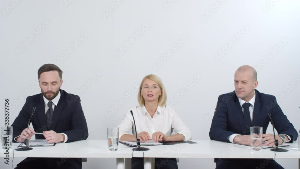 Professional businesswoman sitting between male colleagues and speaking into microphone while answering questions during press conference Professional businesswoman sitting between male colleagues and speaking into microphone while answering questions during press conference