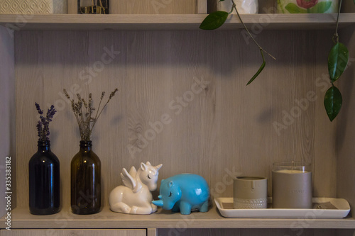 Decorations for the house stood on a shelf of the cupboard (bottles with dried flowers, piggy banks in the form of a white rhino and blue unicorn, candle-sticks) in warm beige colors.	