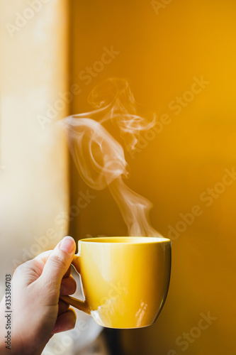 Yellow cup with hot steaming tea drink in hand near window with bright yellow wall background