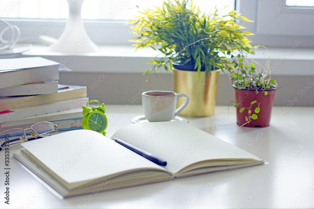 Workplace at home.Written white table by the window with books, a notebook for text, a flower, an alarm clock, a lamp, a cup of coffee. Home office concept, freelance, home schooling Workplace at home.Written white table by the window with books, a notebook for text, a flower, an alarm clock, a lamp, a cup of coffee. Home office concept, freelance, home schooling