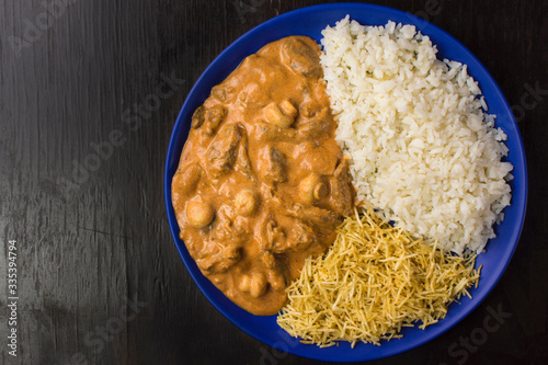 Brazilian tenderloin stroganoff with rice and potato sticks in a blue plate isolated in dark wood background  seen from above