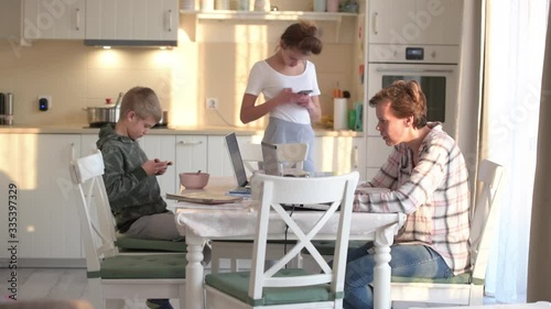 Mother, son and daughter looking, scrolling at their digital devices, smartphone and laptop in the kitchen separately, no communication
