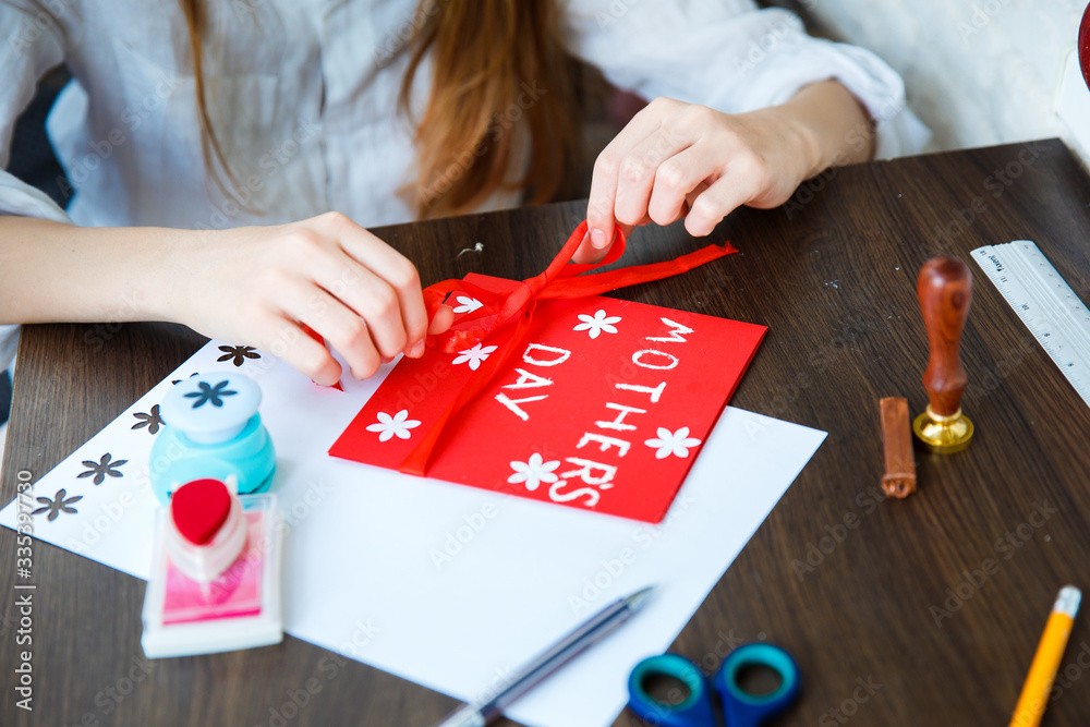 Obraz premium Child holds a card with flowers in his hands. Glue, scissors on a wooden table. Paper flower craft for kids. Child art project.