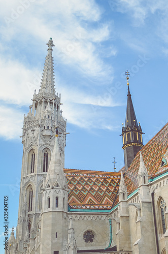 The spire of the famous Matthias Church in Budapest, Hungary. Roman Catholic church built in the Gothic style. Orange colored tile roof. Blue sky and white clouds above. Vertical photo with filter