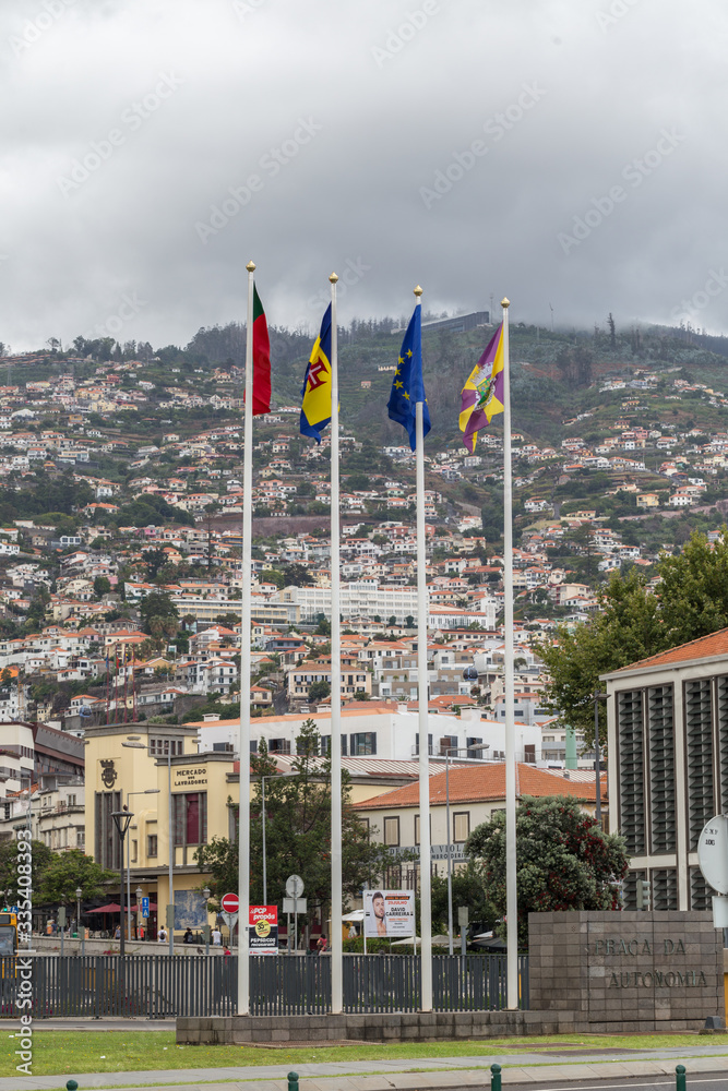 Flags of Portugal, Madeira, the European Union and Funchal against the background of the city buildings of Funchal.