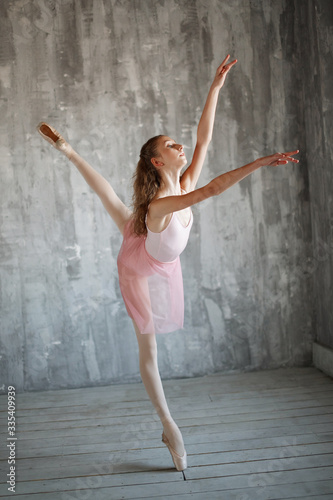 Beautiful female flexiable ballet dancer doing exercise on a gray background. Adorable ballerina is wearing a pink leotard and skirt, beige stockings, pointe shoes.