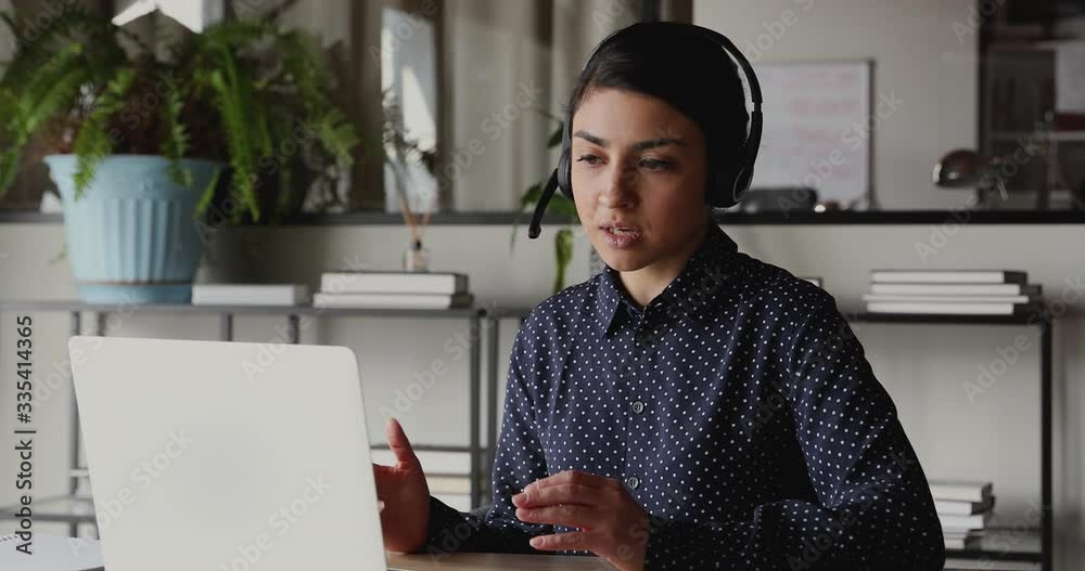 Confident indian female web coach, online teacher or customer support service operator wearing headset speaking to webcam looking at laptop. Ethnic business woman making conference video call concept.