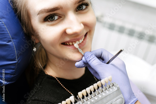 Dentist checking on female patient teeth color, using tooth enamel scale, dental office interior. The blonde smiles in the dental chair, looking forward to the whitening procedure
