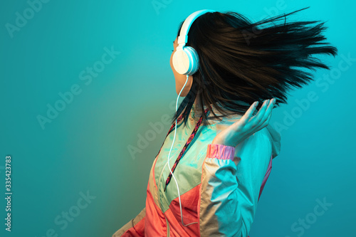 Happy young woman in 90s style sport outfit with white headphones dance in neon lights at studio.
