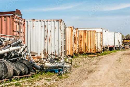 Old rusting industrial shipping containers stored in a field along a dirt road - with salvage scrap and old tires.