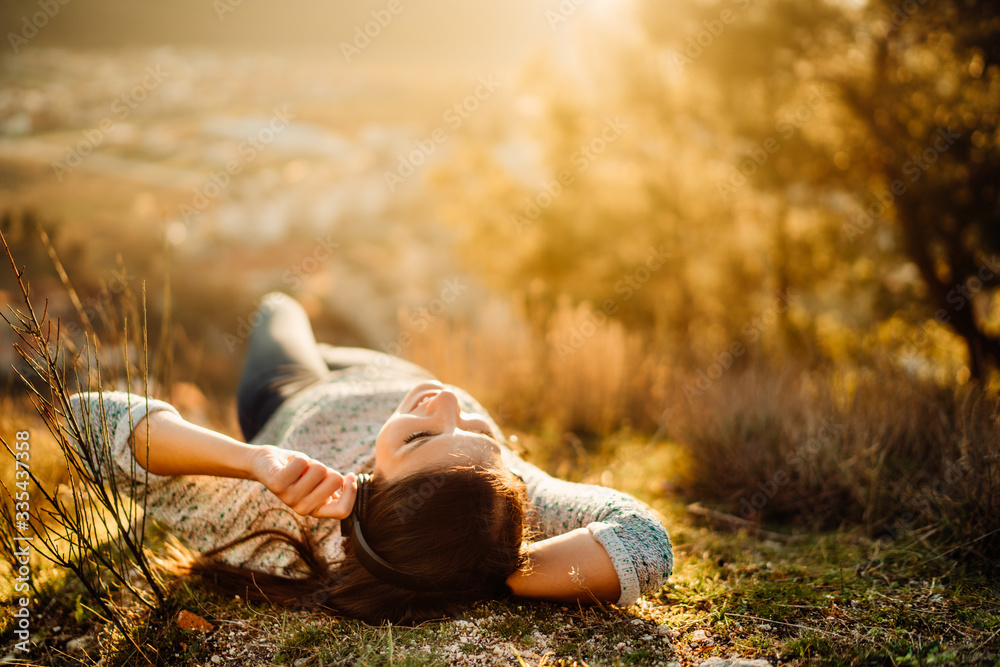 Carefree happy woman lying on green grass meadow on top of mountain enjoying sun on her face.Enjoying nature sunset.Freedom.Relaxing in mountains at sunrise.Sunshine.Daydreaming.Listening to music - obrazy, fototapety, plakaty 