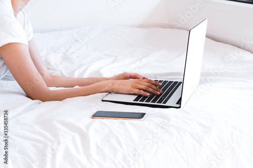 home office in a bed. young woman wearing white shirt and blue jeans and typing on a white and black laptop keyboard on white crumpled sheets