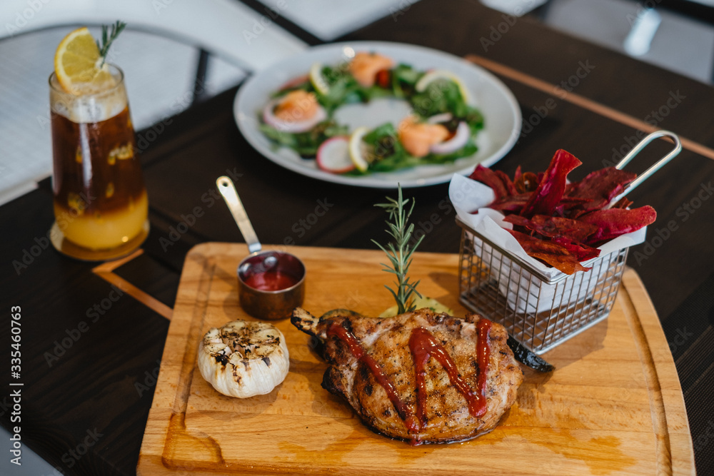 grilled pork chop with raspberry sauce and grilled garlic, potato, pumpkin vegetable on wooden board served with potato chips in basket. with salmon salad and orange mix cold brew coffee on table. grilled pork chop with raspberry sauce and grilled garlic, potato, pumpkin vegetable on wooden board served with potato chips in basket. with salmon salad and orange mix cold brew coffee on table.