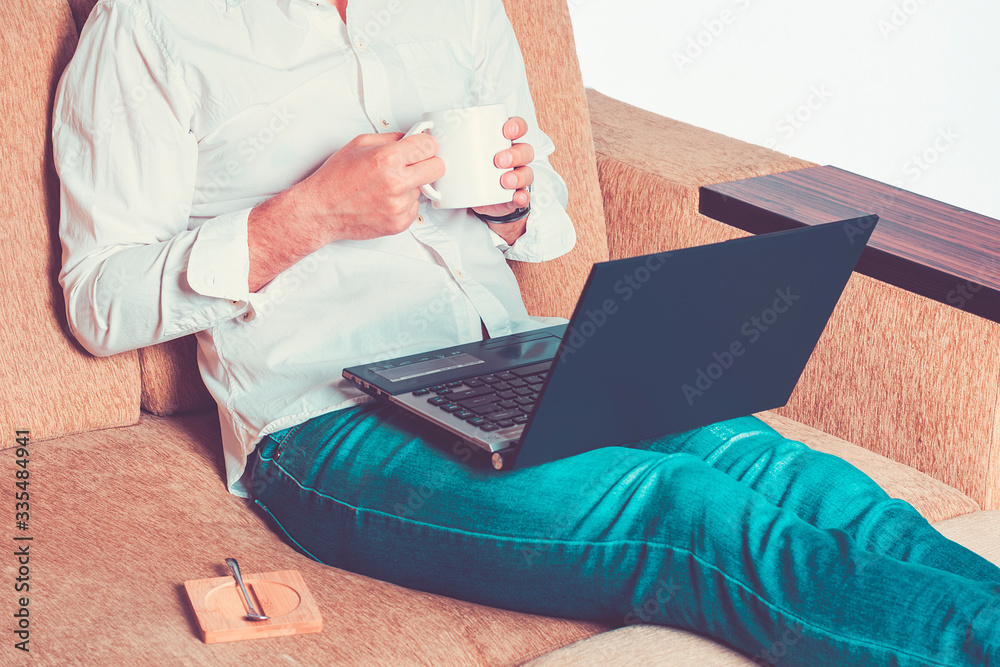 Naklejka premium young man in white shirt and blue jeans hold cup of tea and working on laptop, writes in notebook in home office sitting on cozy beige sofa Stay at home, online work, distance learning concept