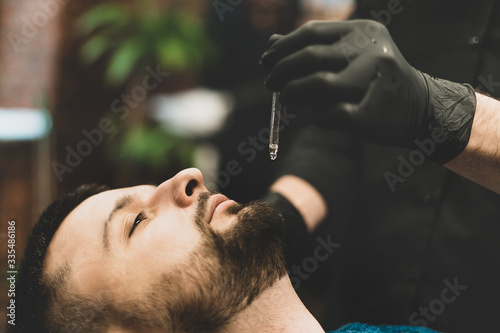 Barbershop. The client in the master’s chair in the barbershop, the barber applies oil and cosmetics to the client’s beard. Male beauty shop. Healthy lifestyle and beauty.