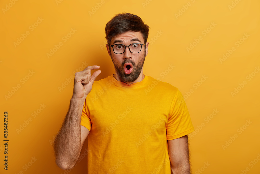 Deeply impressed bearded man shocked by very tiny size, measures something small, gasps from amazement, has unexpected reaction on decreased price, stares through glasses, wears yellow t shirt Deeply impressed bearded man shocked by very tiny size, measures something small, gasps from amazement, has unexpected reaction on decreased price, stares through glasses, wears yellow t shirt