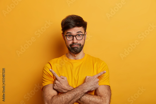 Perplexed unsure European man crosses hands and points sideways, chooses between two objects, should to decide, faces complicated choice, indicates good products, asks your opinion or advice