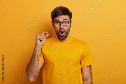 Deeply impressed bearded man shocked by very tiny size, measures something small, gasps from amazement, has unexpected reaction on decreased price, stares through glasses, wears yellow t shirt photo