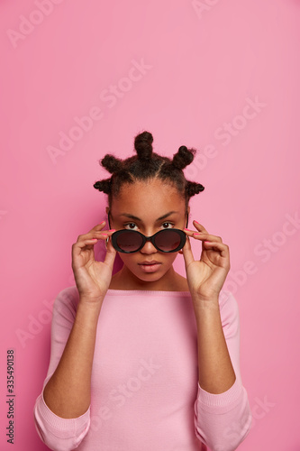 Fashion model with bun hairstyle looks seriously through sunglasses, wears rosy jumper, stands alone, going to have walk during sunny day, poses against pink background, copy space above for text