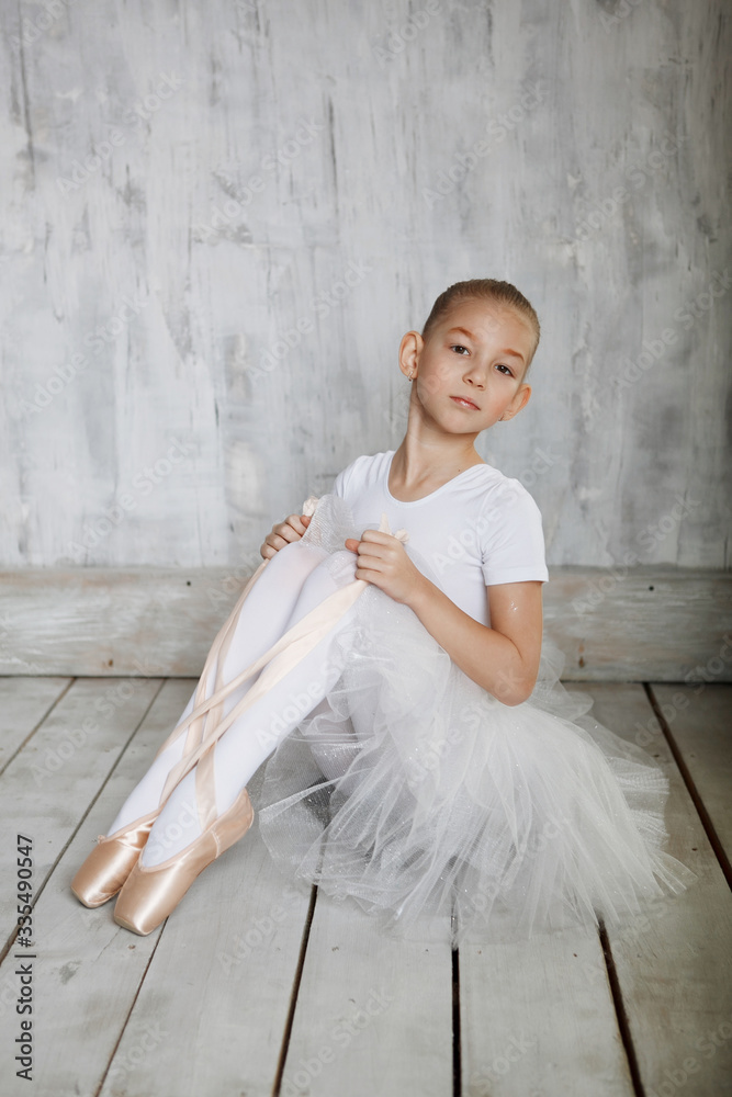 Naklejka premium A little adorable young ballerina in white dress trying on her point shoes sitting on woody floor and gray studio background
