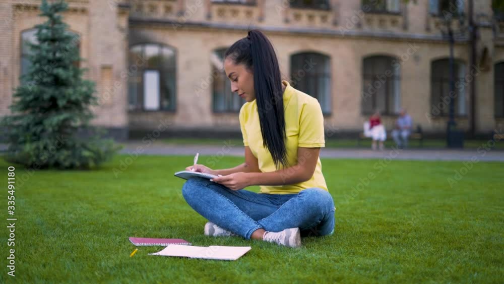 Focused girl student doing homework sitting on grass on campus takes notes in a notebook. 4K Focused girl student doing homework sitting on grass on campus takes notes in a notebook. 4K