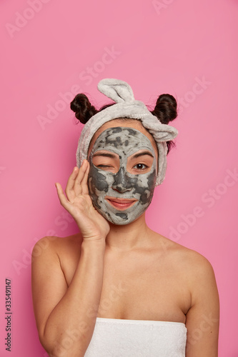 Studio shot of lovely young ethnic woman applies face mask with mud  winks eye  touches cheeks  enjoys cosmetology procedure  follows advice of beautician  cares about body skin and complexion