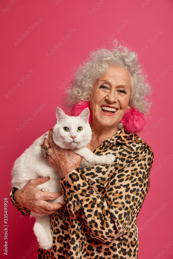 Indoor shot of happy wrinkled granny petting her white cute cat, being old friends, dressed in fashionable clothes, amuse together on retirement. Old people, pets, happiness, leisure concept Indoor shot of happy wrinkled granny petting her white cute cat, being old friends, dressed in fashionable clothes, amuse together on retirement. Old people, pets, happiness, leisure concept