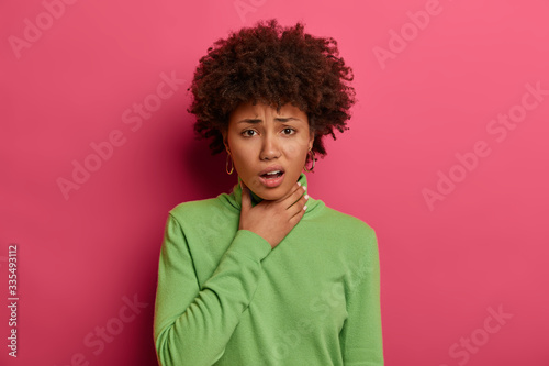 Sick dark skinned young woman feels unwell, suffers from sore throat, has suffocation and cough, frowns face, wears green turtleneck, feels pain after catching cold, isolated on pink background. © wayhome.studio 