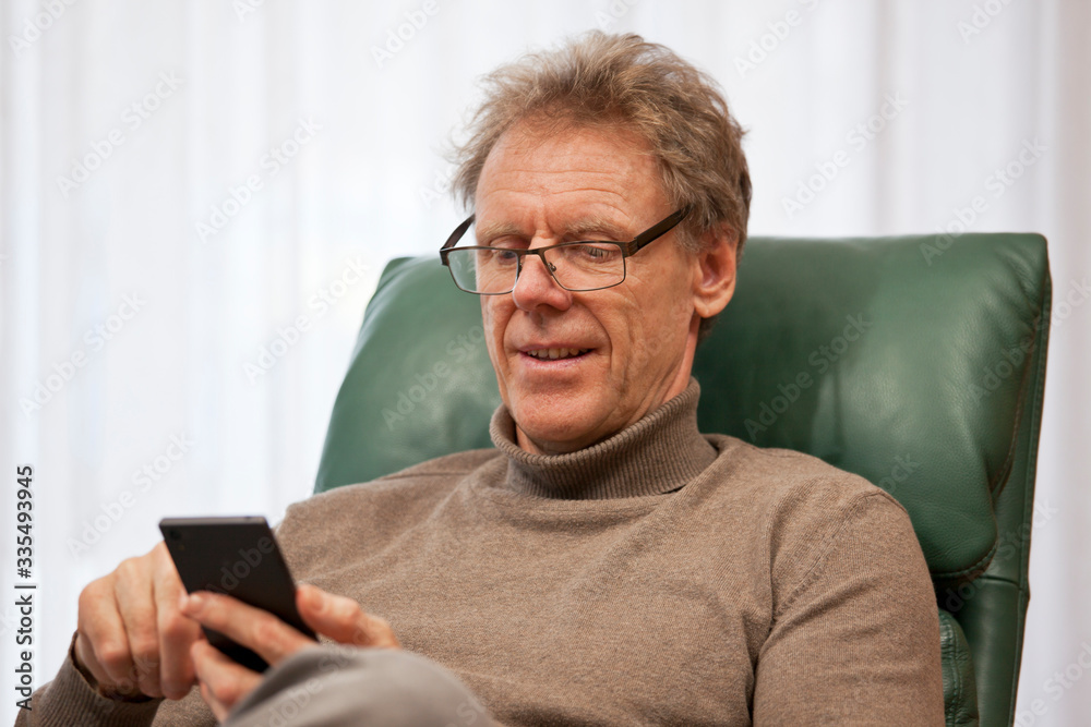 Senior man sitting in a chair in a bright living room looking at a cell phone at home Senior man sitting in a chair in a bright living room looking at a cell phone at home