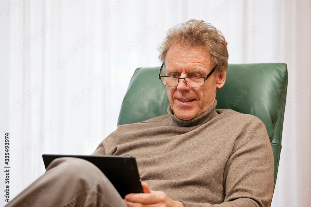 Senior man sitting in a chair in a bright living room looking at a notebook or tablet at home Senior man sitting in a chair in a bright living room looking at a notebook or tablet at home