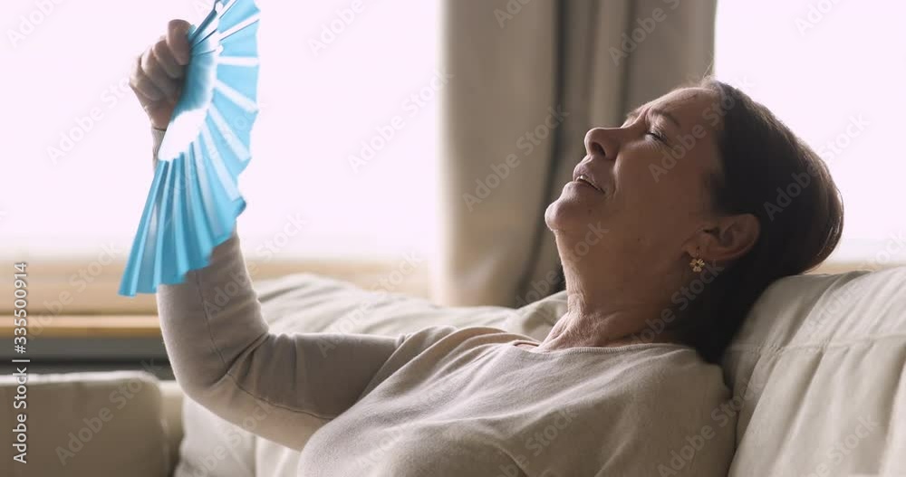 Side view head shot overheated middle aged mature woman using paper fan, suffering from high temperature indoors. Stressed senior older grandmother feeling hot without air conditioning system at home. Side view head shot overheated middle aged mature woman using paper fan, suffering from high temperature indoors. Stressed senior older grandmother feeling hot without air conditioning system at home.