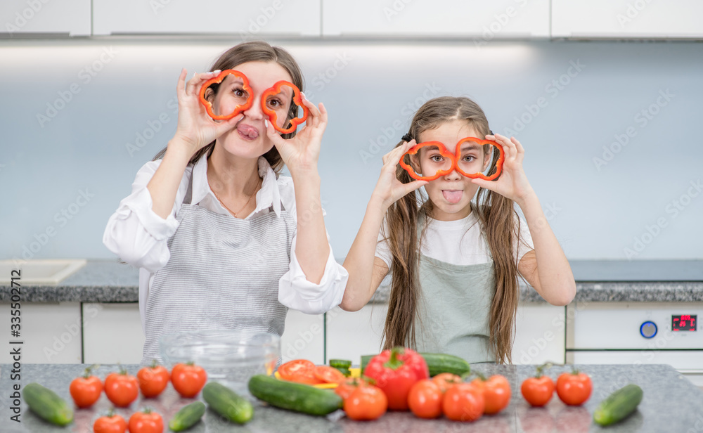 Happy family at home. Mom and her daughter preparing healthy food and having fun, holds a pepper in front of their eyes like a glasses Happy family at home. Mom and her daughter preparing healthy food and having fun, holds a pepper in front of their eyes like a glasses